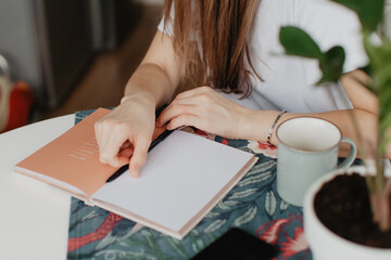 A young woman is sitting at a table in the kitchen with a cup of coffee and a diary