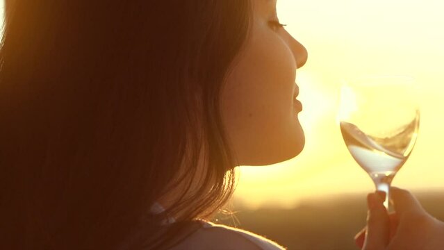 Beauty Young woman drinking white wine and enjoying sunset view on a terrace. Beautiful Caucasian girl holding glass of wine outdoors. Wine tasting. Enjoying of wine over sun light. Slow motion