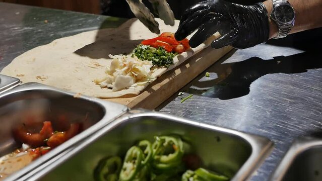 the hand of the cook in a black glove puts fresh pieces of tomato with culinary tongs into a thin pita bread with other vegetables. The process of making lula kebab and shish kebab.
