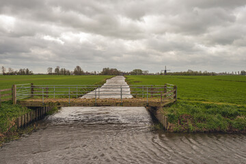 Dutch polder landscape with a simple bridge between the meadows in the foreground and a mill built in 1581 in the background. The photo was taken on a cloudy day at the beginning of the spring season.