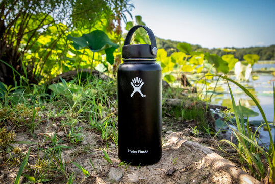 Fayette, MO United States of America - September 1st, 2021 : Hydroflask black insulated water bottle, sitting on the shore of a lake, surrounded by vegetation and lily pads in the shade.  