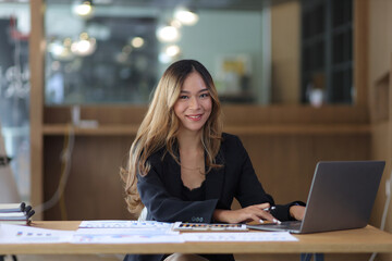 Fototapeta premium Portrait of a business woman working or secretary with laptop and financial documents on the desk in the office. Looking at camera.