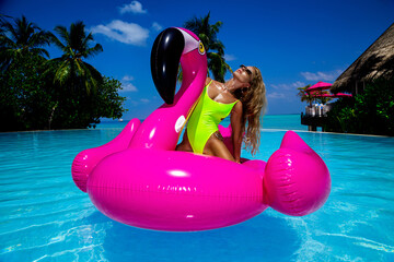 Beautiful sexy tanned woman on pink flamingo in pool. Young glamour girl in swimsuit on Maldives...