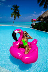 Beautiful sexy tanned woman on pink flamingo in pool. Young glamour girl in swimsuit on Maldives...
