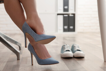 Comfortable sneakers on floor near woman wearing stylish high heeled shoes in office, closeup