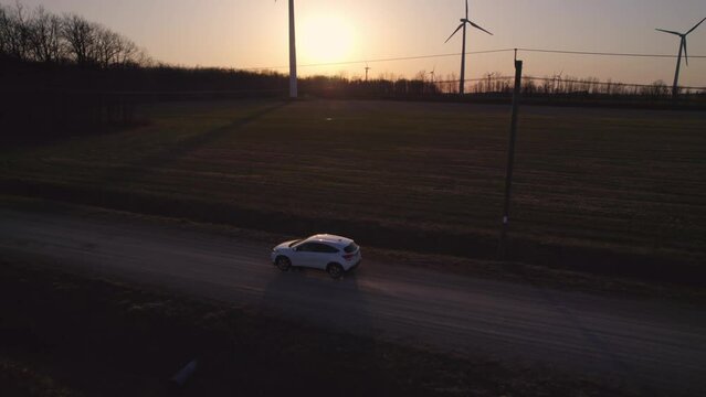 Aerial shot of car SUV driving at the golden hour by rural road. Vehicle travels near beautiful farming fields and electric wind turbines surrounded by trees. Alternative energy, agriculture concept.