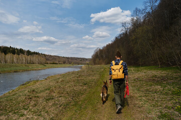 Man wears backpack walks with German Shepherd along path river. Owner and dog in park, rear view. Nature of the Moscow region, the Moskva River in the countryside on a warm summer sunny day.