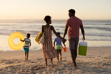 Rear view of african american parents carrying blankets and cooler walking with children at beach