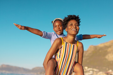 African american young woman piggybacking playful daughter stretching arms against clear blue sky