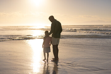 Full length of african american young man with son standing at shore against sky during sunset