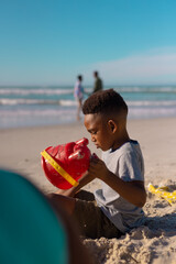Side view of african american boy holding pail playing with sand on beach against sea and blue sky
