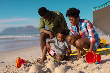 African american young parents looking at son playing with sand at beach against sky on sunny day