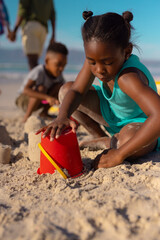 African american girl making sand castle with pail at beach against sky with brother in background