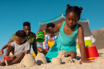 Low angle view of african american girl making sand castle against sky with family in background