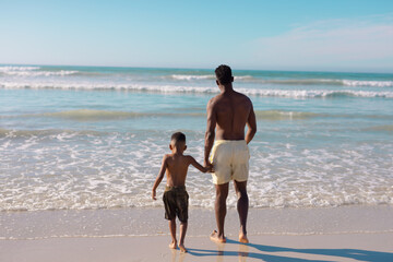 Rear view of african american shirtless young man holding son's hands walking at beach against sky