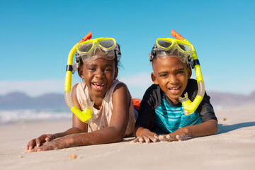 Portrait of happy african american boy and girl wearing snorkels and masks lying on sand against sky - obrazy, fototapety, plakaty