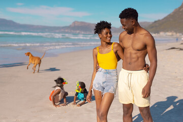 Smiling african american young couple walking while children and dog playing at beach against sky