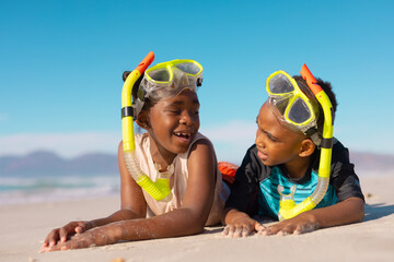 African american boy and girl wearing snorkels and masks talking while lying on sand against sky