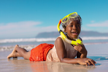 Portrait of happy african american wet girl wearing mask and snorkel lying at shore against blue sky - obrazy, fototapety, plakaty