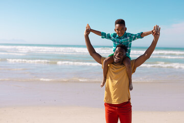 Cheerful african american young man carrying son on shoulders standing against sea and blue sky