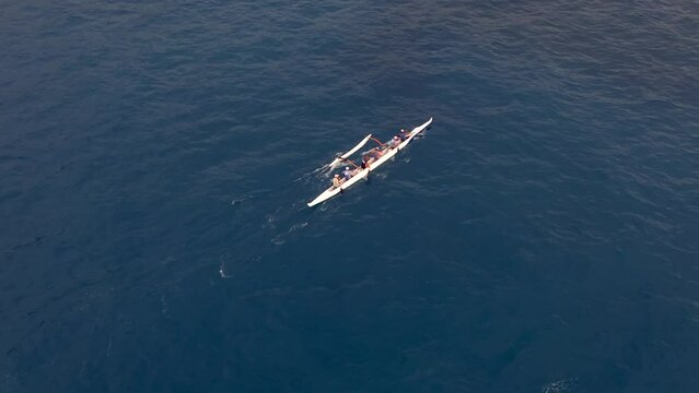 A rowing team paddles a Hawaiian outrigger canoe along the ocean in synchrony - aerial bird's eye view