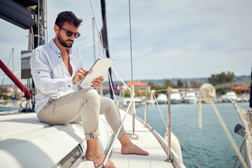 A young businessman is sitting on his yacht and doing some job on his tablet while riding in the dock on the seaside. Summer, sea, vacation