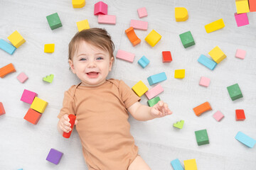 cute smiling baby girl in brown bodysuit laying on floor around colorful wooden blocks. top view