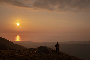 A man standing beside a wild camping tent at sunset in Snowdonia UK