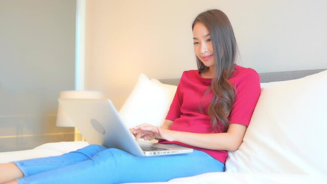 While sitting on a bed, a young casually dressed woman works on her laptop.