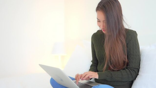 Close-up of a casually dressed young woman sitting on a bed as she works on her laptop. Title space