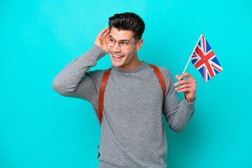 Young caucasian man holding an United Kingdom flag isolated on blue background listening to something by putting hand on the ear