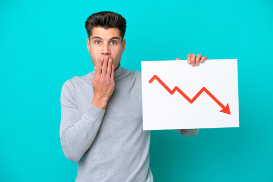 Young handsome caucasian man isolated on blue bakcground holding a sign with a decreasing statistics arrow symbol with surprised expression