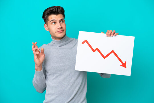 Young handsome caucasian man isolated on blue bakcground holding a sign with a decreasing statistics arrow symbol with fingers crossing
