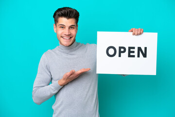 Young handsome caucasian man isolated on blue bakcground holding a placard with text OPEN and  pointing it