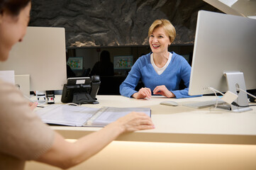 Beautiful pleasant Caucasian female travel agent talking to her client, showing catalog with tours...