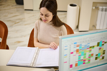 Beautiful young European woman leafing through a catalog while sitting at a table in the waiting room of an administrative building with monitor turned on and schedule chart in the blurred foreground