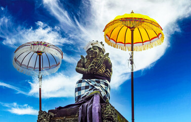 Balinese hindu mythological lord shiva stone figure between two traditional ceremonial umbrellas (tedung), blue sky white clouds - Bali, Ubud