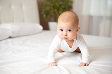 Portrait of a cute baby girl of 5 months in a white bodysuit on a white bed in the bedroom. Space for text. Child lifestyle