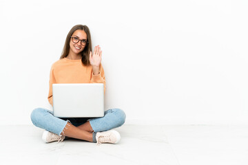Young woman with a laptop sitting on the floor saluting with hand with happy expression