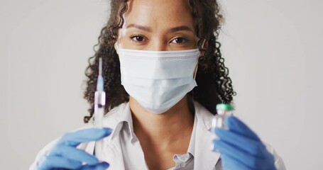 Video of portrait of biracial female doctor with face mask holding vaccine vial on white background - Powered by Adobe