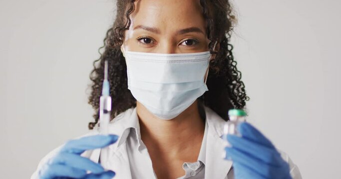 Video of portrait of biracial female doctor with face mask holding vaccine vial on white background