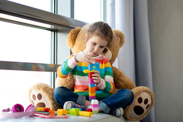 A cute little girl plays construction set. Childhood. Development. She sits near a window with a large teddy bear. The girl is dressed in a colored sweater and jeans.