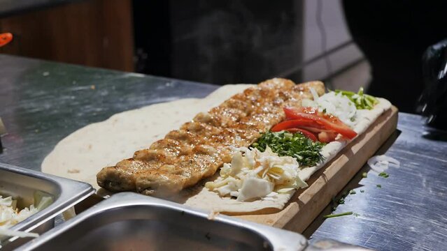 serving ready-made hot lula kebab on skewers to pita bread with chopped pieces of fresh vegetables and herbs in a restaurant. The cook takes the skewers out of the barbecue