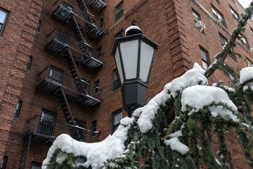 Street Light next to a Snow Covered Evergreen Tree in front of an Old Brick Apartment Building in...