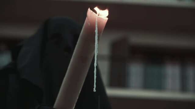 Close-up shot of the melting candle and in the background the covered face of a worshipper in a black tunic. Holy Week in Spain.