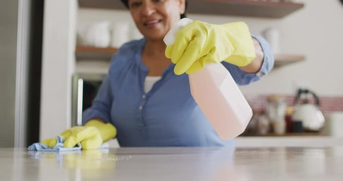 Smiling senior biracial woman wearing gloves and cleaning table in kitchen alone