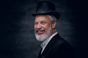 Shot of joyful senior man dressed in black suit with hat looking at camera against dark background.