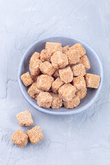 Cubes of brown sugar or cane sugar in bowl and on gray background, vertical, top view
