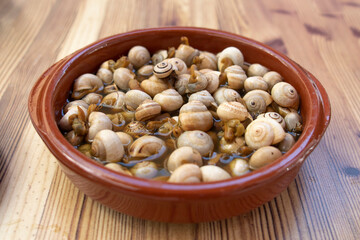A terracotta bowl with snails in broth, on a wooden table. Spanish tapas highly demanded. Spanish...
