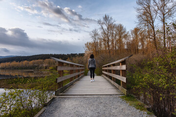 Fototapeta na wymiar Woman Walking on a Wooden Path with green trees in Shoreline Trail, Port Moody, Greater Vancouver, British Columbia, Canada. Trail in a Modern City during a Cloudy Sunset.
